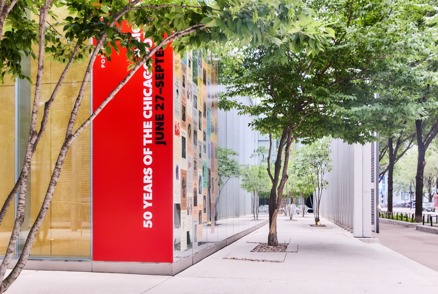 Outdoor view of a building entrance with a tall red banner reading "50 Years of the Chicago Poetry Center" alongside a collage of historic event posters, framed by leafy green trees along a city sidewalk.