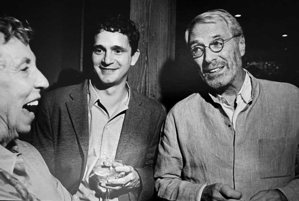 Black-and-white photo of three men sharing a laugh at a social gathering, the center figure holding a drink, in what appears to be a Poetry Center event from past decades.