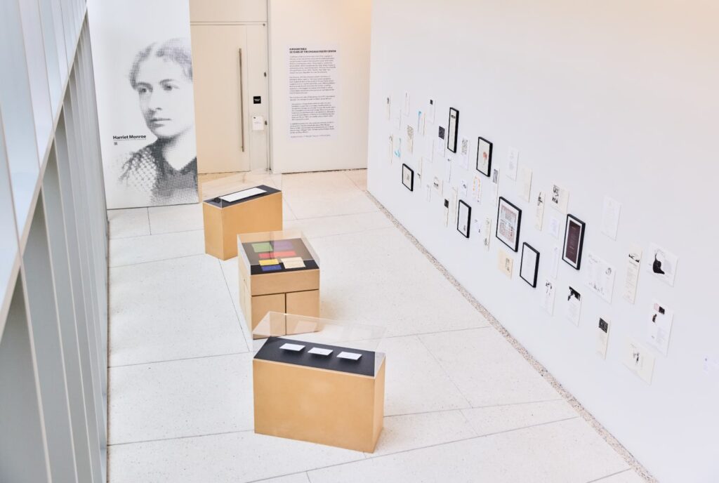 Interior view of the "A Bigger Table: 50 Years of the Chicago Poetry Center" museum exhibition, featuring a large halftone portrait of Harriet Monroe, display cases with archival materials, and a wall of historic documents and broadsides.