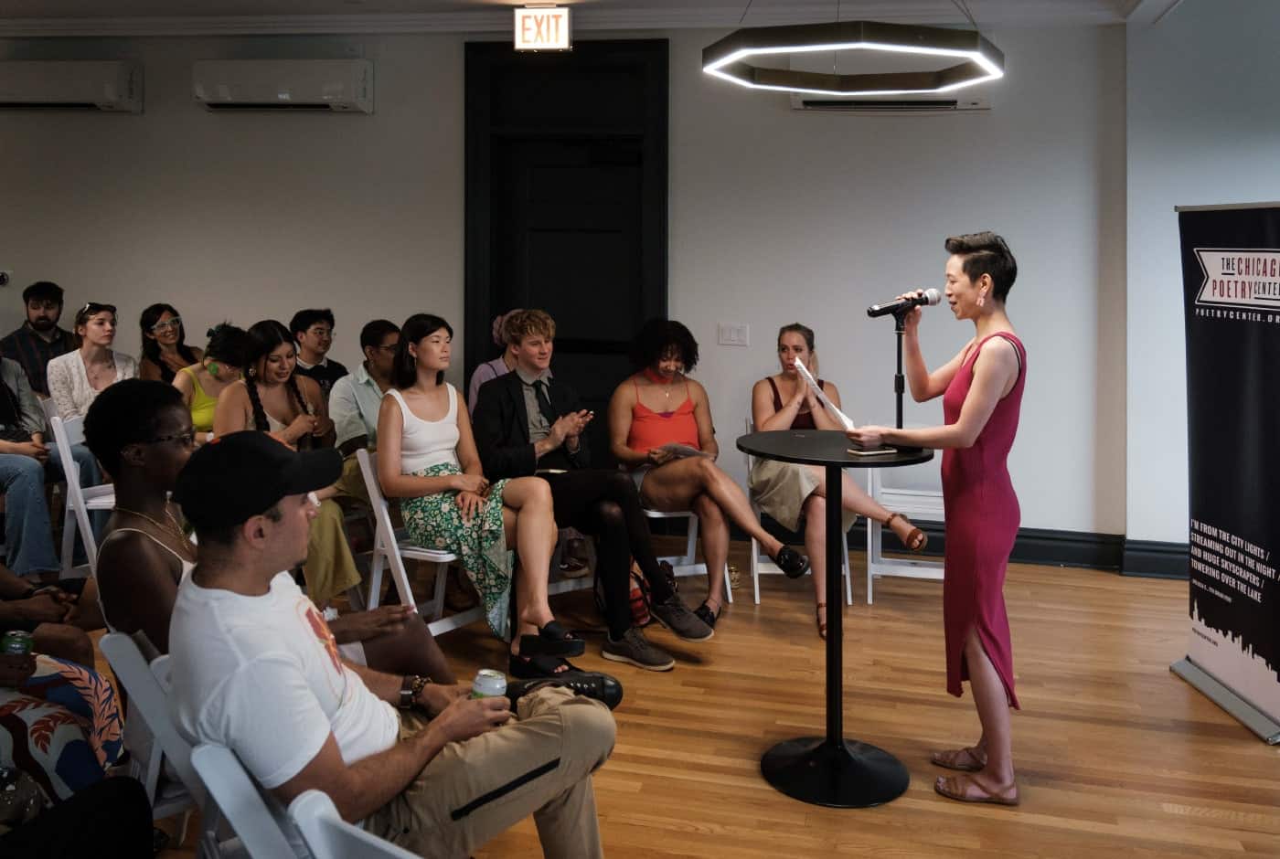 A poet in a magenta dress reads at a microphone to a seated audience in a warmly lit event space, with a Chicago Poetry Center banner beside her.