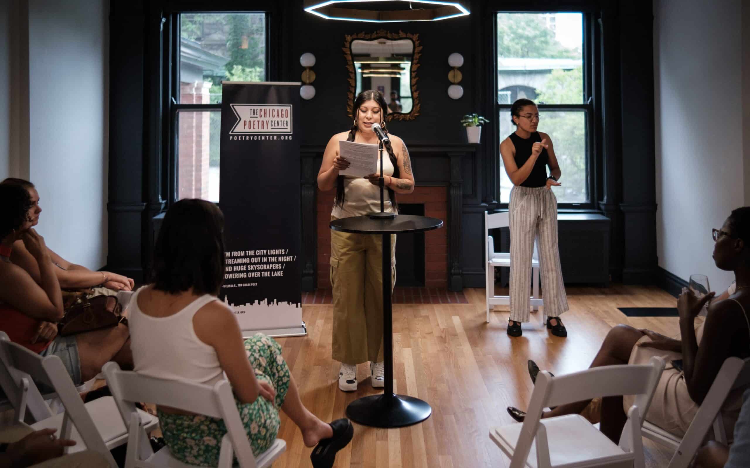 A poet reads from a paper at a microphone in an elegant room with dark trim and large windows, while an ASL interpreter signs nearby for the audience—a Blue Hour reading event.