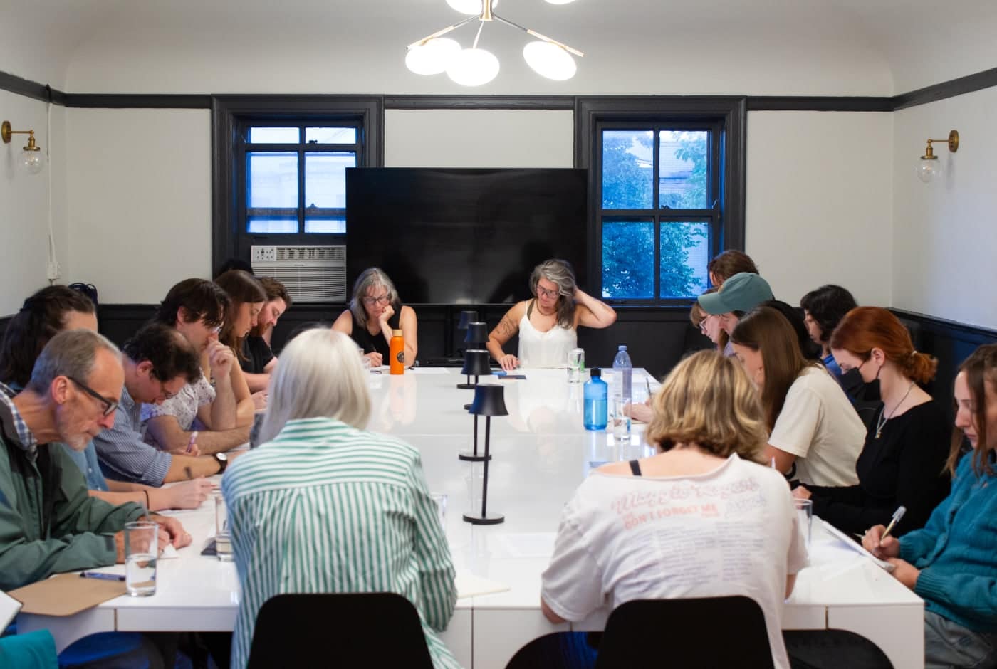 Participants sit around a long table during a Blue Hour generative writing workshop, bent over their notebooks in concentration, with dusky blue light visible through the windows.