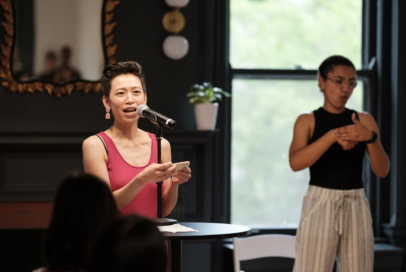 A poet in a pink tank top reads from her phone at a microphone while an ASL interpreter signs beside her in an elegant room with dark-framed windows and natural light.