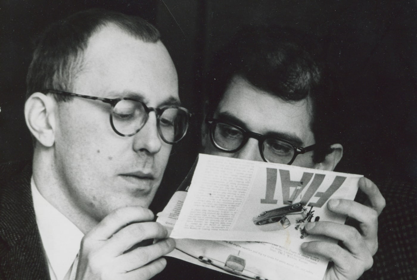 Black-and-white archival photo of two men in glasses examining a magazine together in close conversation. Features Paul Carroll, American poet and the founder of the Poetry Center of Chicago.