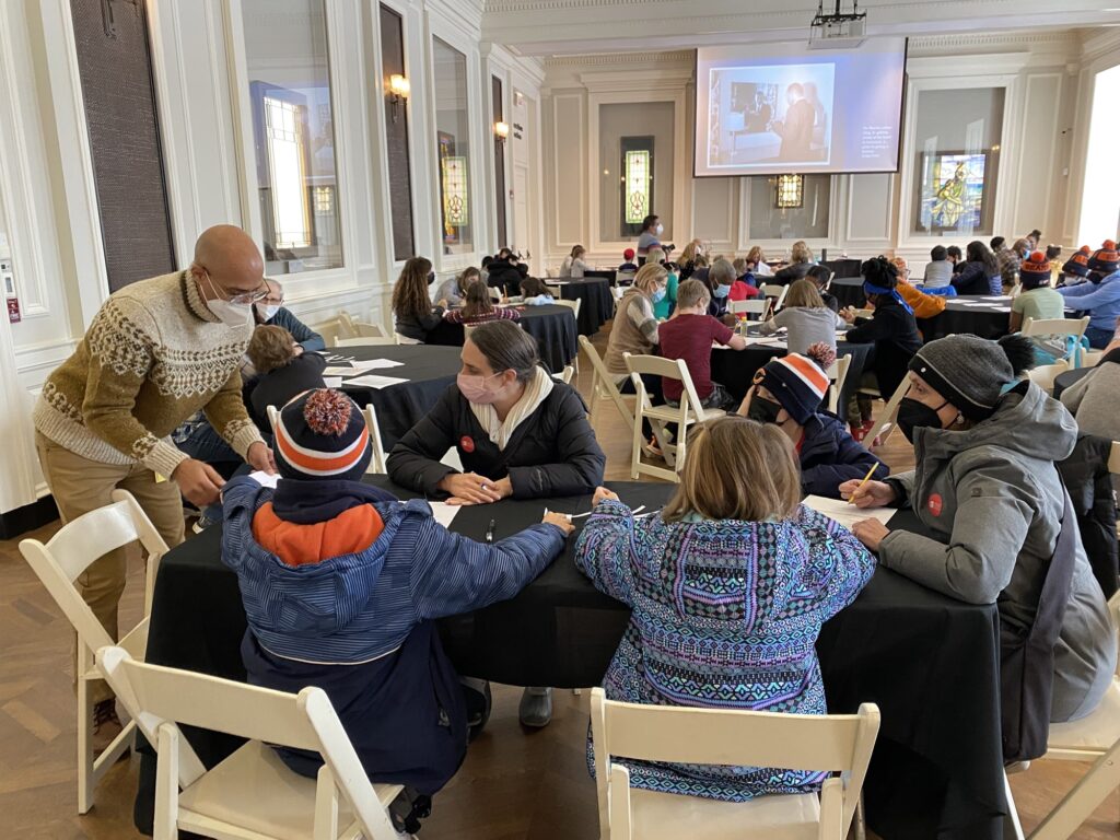 A teaching artist in a patterned sweater assists families at round tables during a poetry workshop in a grand hall with stained glass windows and a presentation screen, with participants wearing winter coats and hats.
