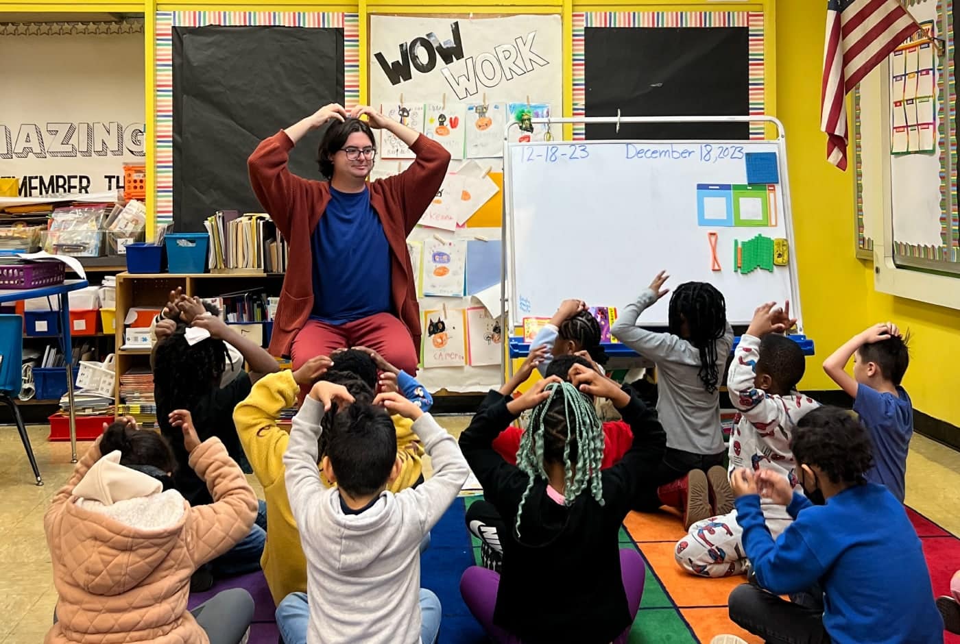 A teaching artist in a rust cardigan leads elementary students in a movement activity, everyone with hands on their heads, as children sit on a colorful rug in a classroom decorated with a "Wow Work" bulletin board.
