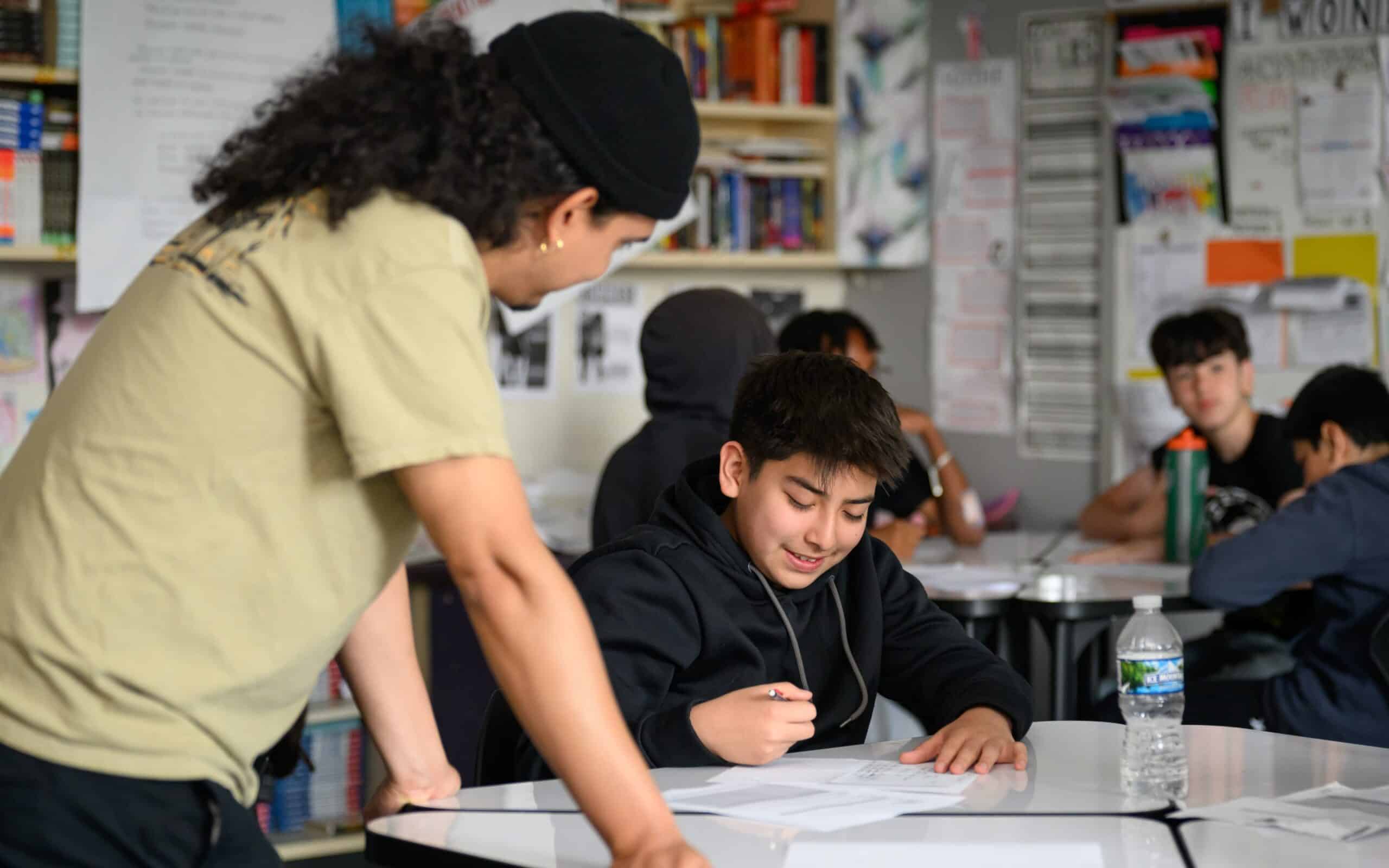 A teaching artist wearing a cap leans over a desk to help a smiling middle school student with their writing, with classmates working in the background.