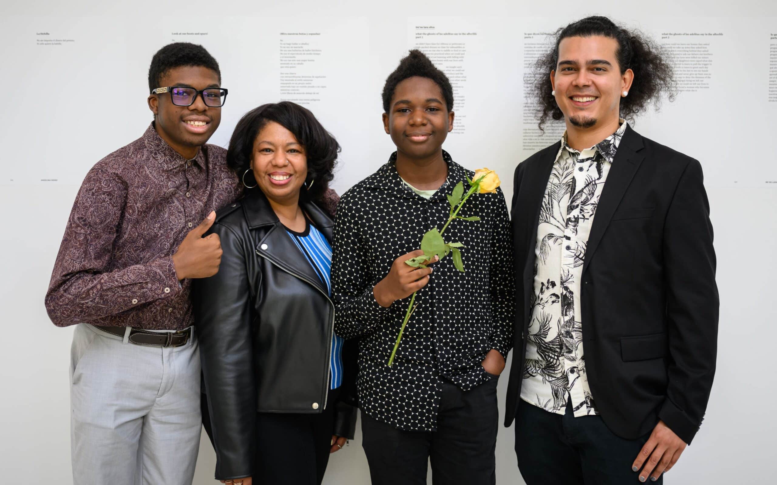 A teaching artist in a suit jacket poses proudly with two student poets—one holding a yellow rose—in front of a wall displaying printed poems.