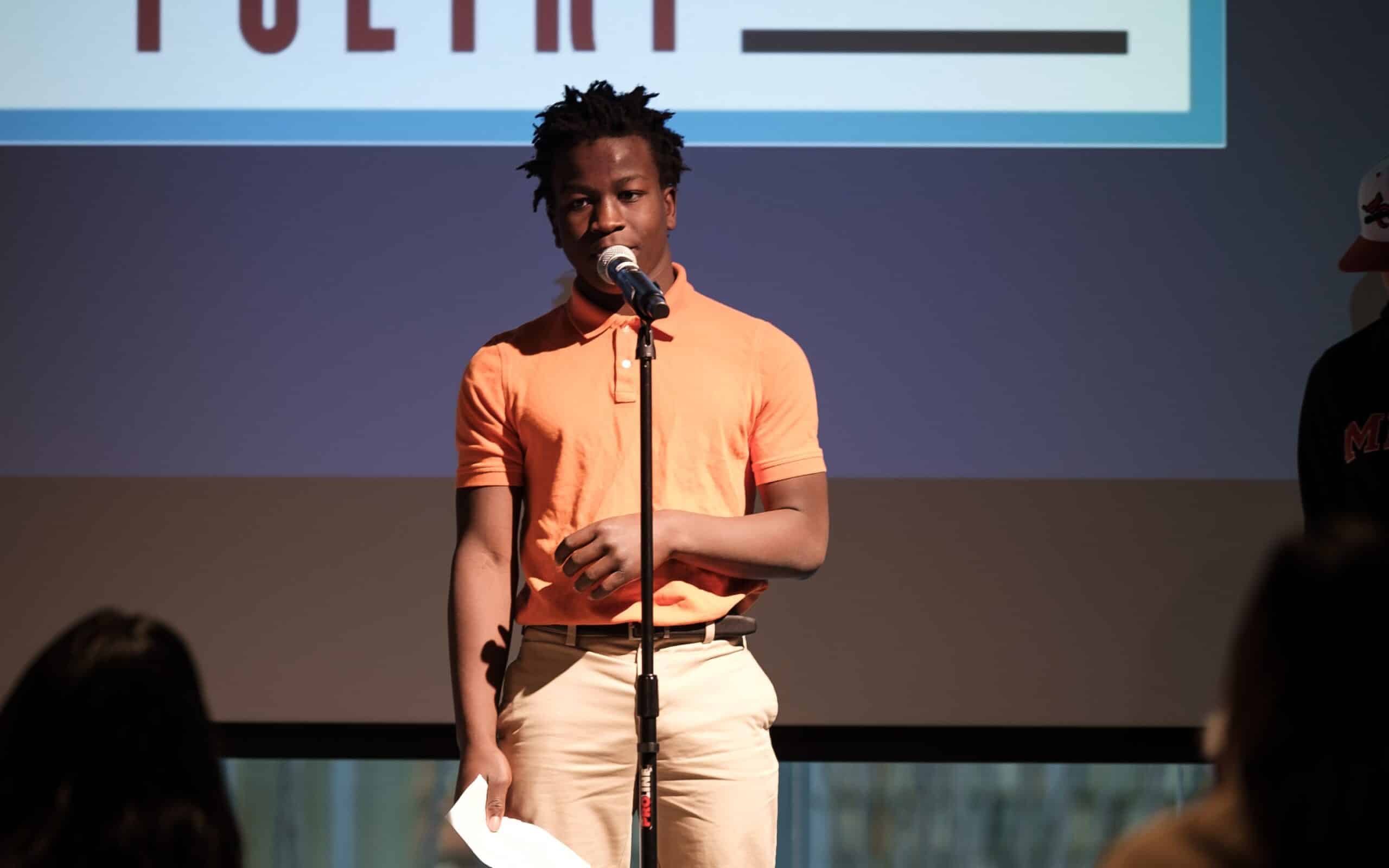 A young man in an orange polo shirt performs at a microphone on stage, holding his poem in one hand, with "POETRY" visible on a screen behind him.