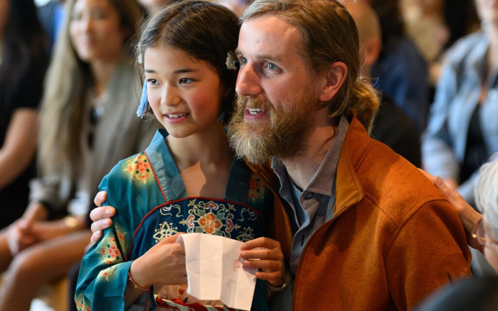 A young girl in a teal embroidered robe holds a folded paper and smiles beside a bearded man in an orange jacket at a poetry reading event, with audience members visible in the background.