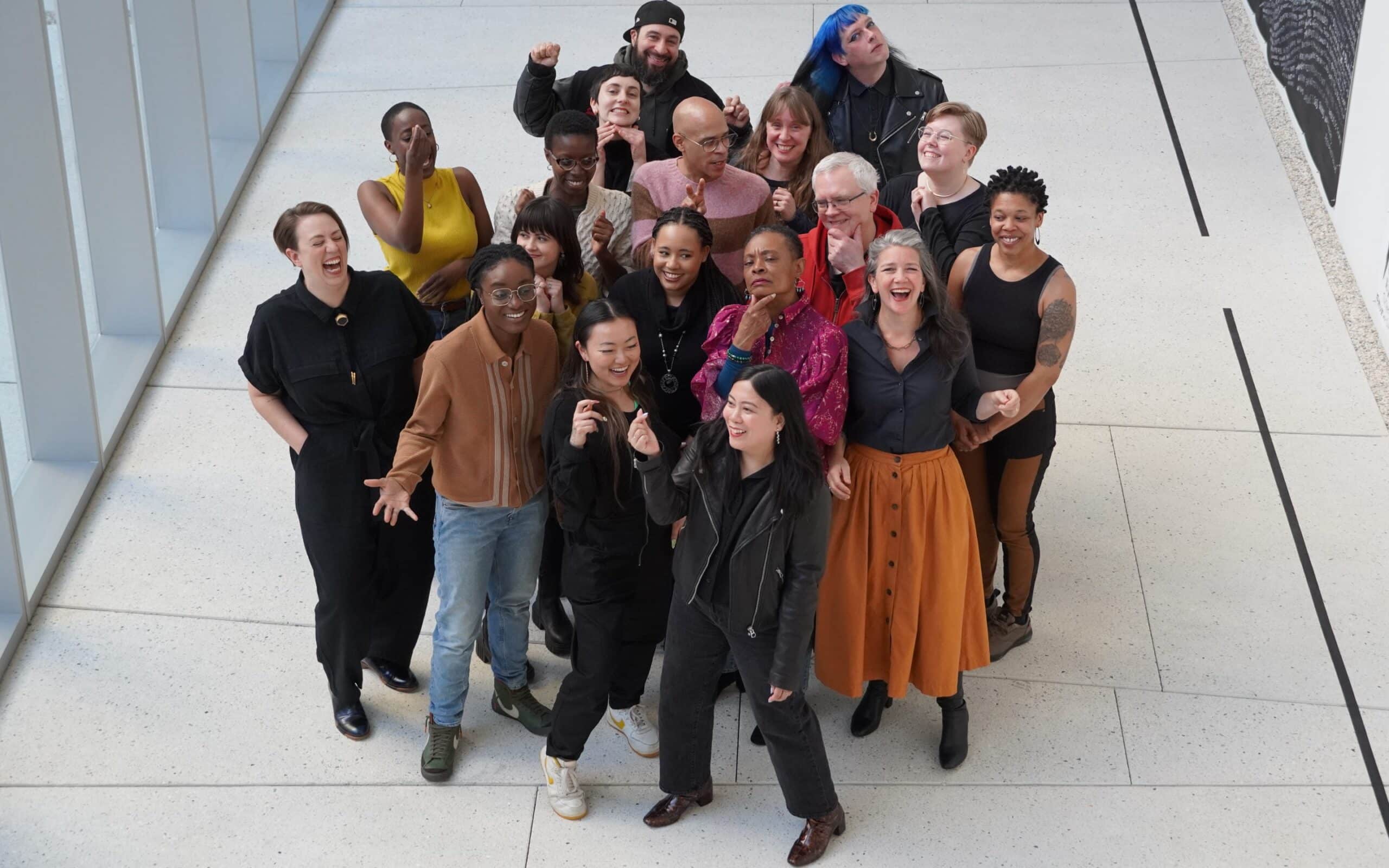 Wide overhead shot of the Chicago Poetry Center team gathered on a museum floor, laughing and posing playfully with raised fists and animated expressions.