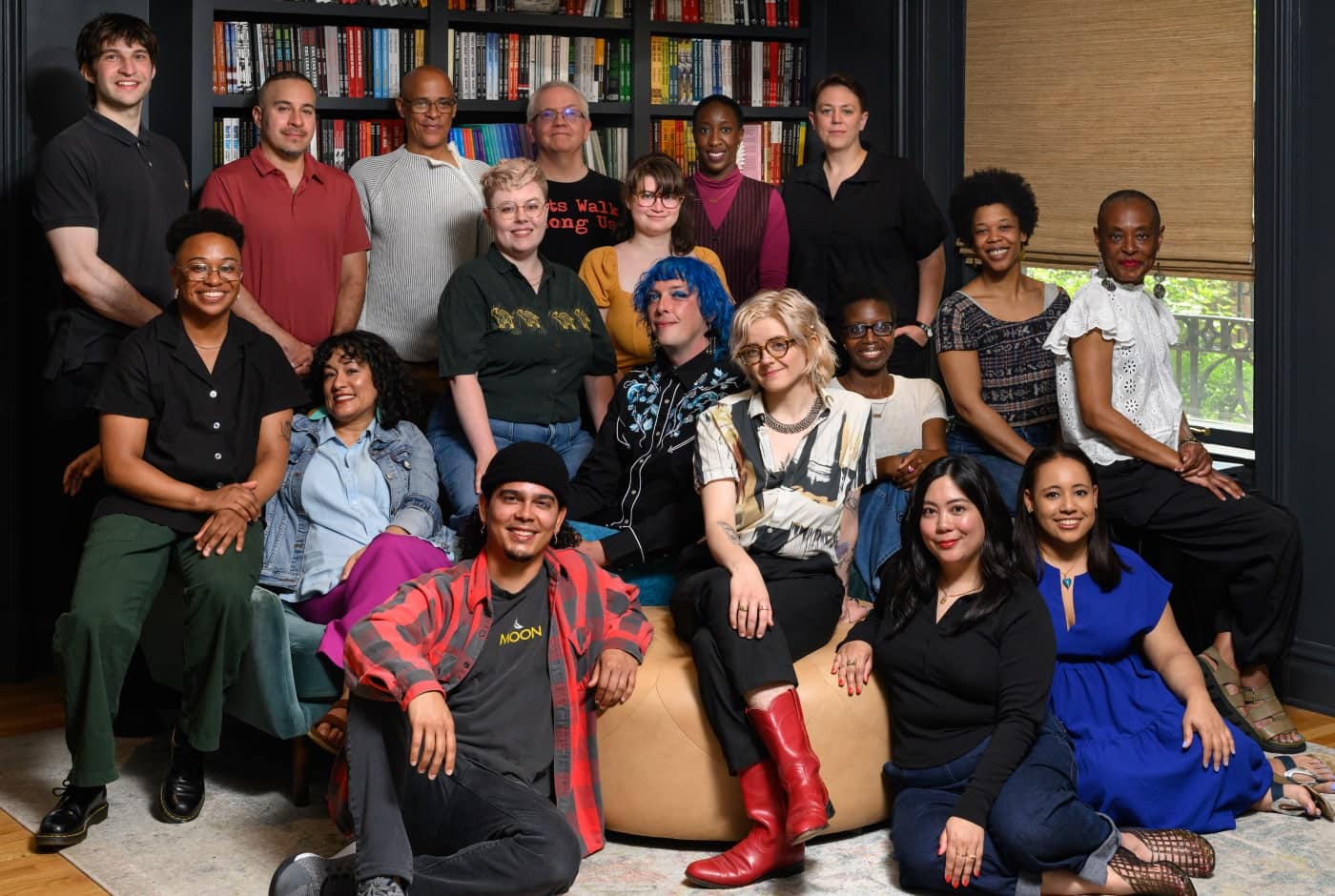 A group portrait of approximately 20 Chicago Poetry Center staff and teaching artists posed in front of a dark bookshelf, with members seated and standing in colorful attire.
