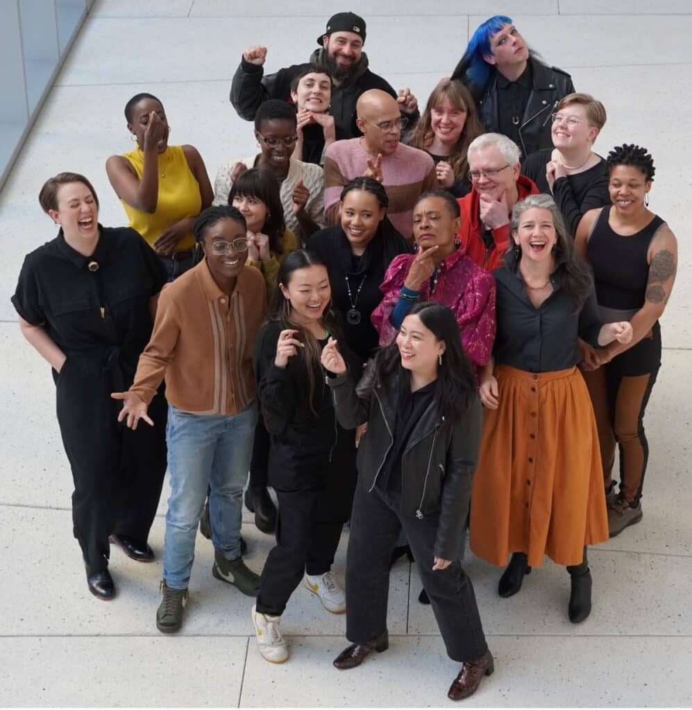 An overhead shot of the Chicago Poetry Center team gathered on a light-colored floor, laughing and striking playful poses with raised fists and expressive gestures