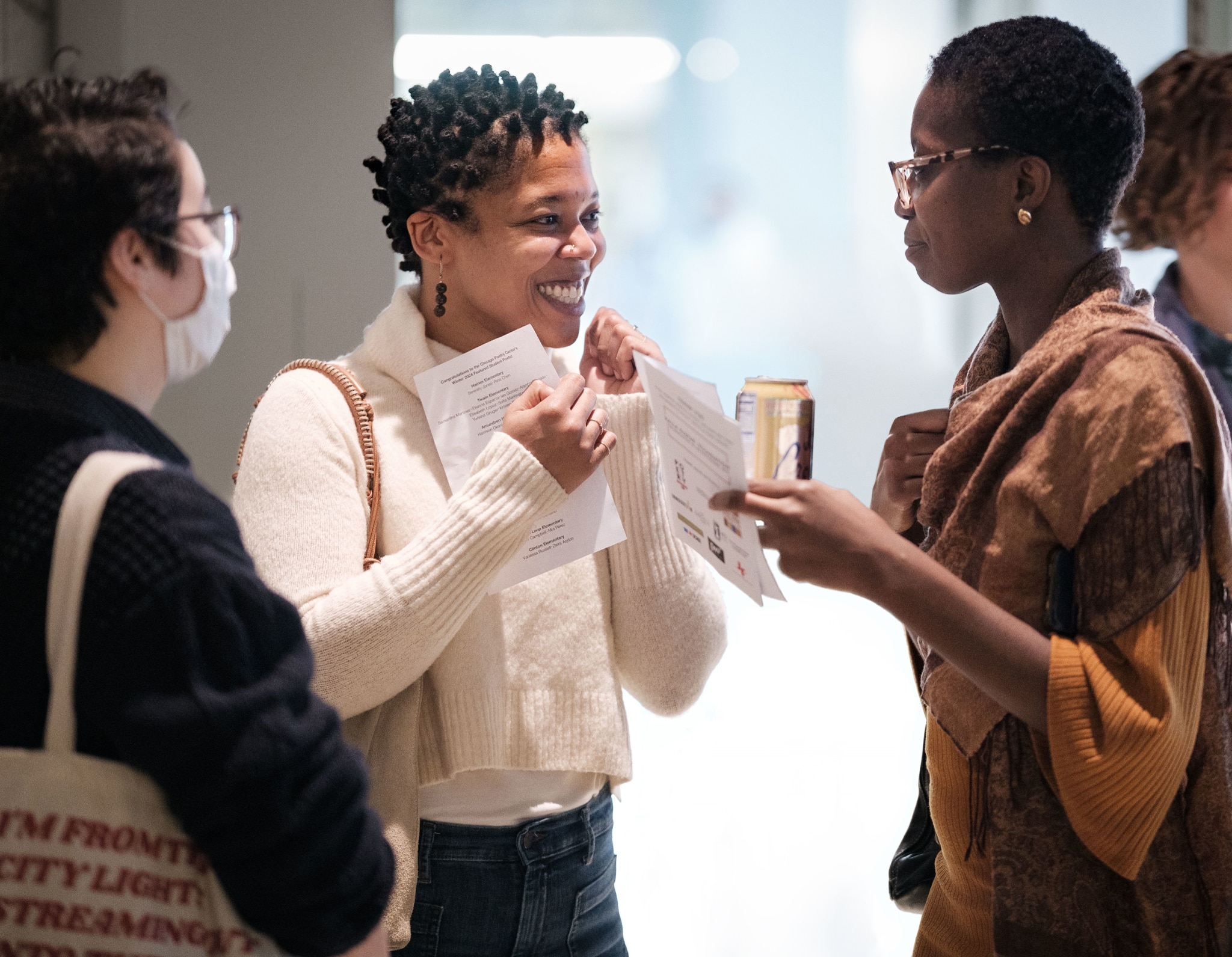 Three women laugh and chat while holding event programs at a poetry gathering, with bright natural light in the background.