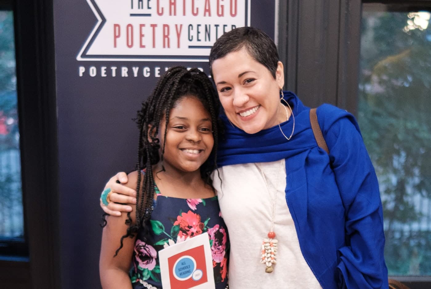 A young student with long braids and a floral dress poses proudly with a teaching artist in front of the Chicago Poetry Center banner, holding an "All Schools Reading" program booklet.