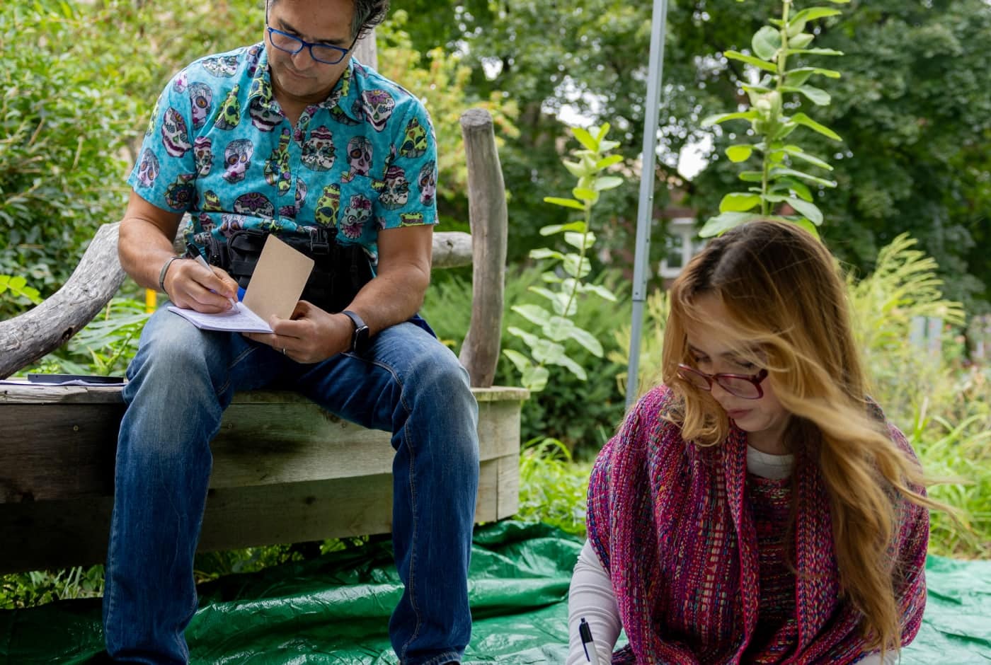 Two people write in notebooks at an outdoor poetry event in a garden, one wearing a colorful skull-print shirt and the other in a pink woven sweater.