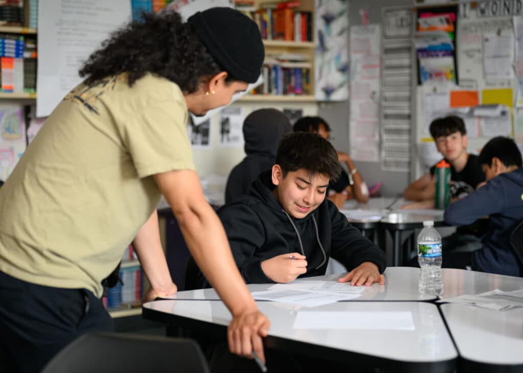 A teaching artist wearing a cap leans over a desk to help a smiling student with their writing in a classroom, while other students work in the background.