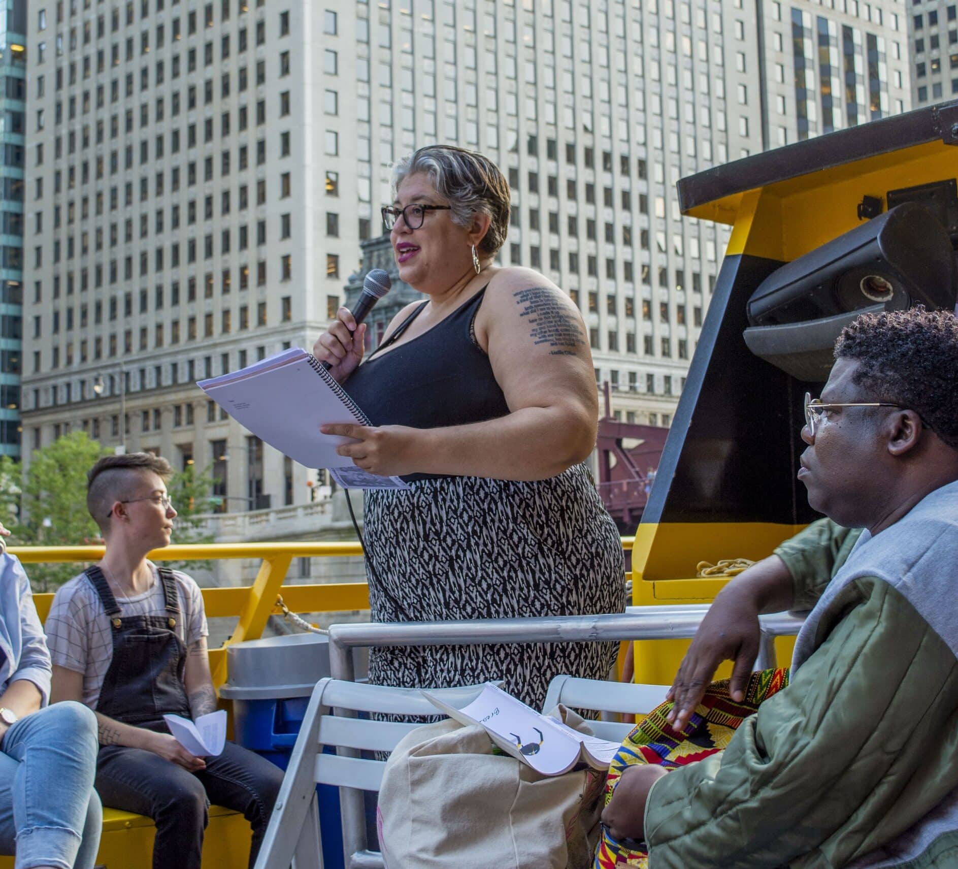 A poet with a text tattoo on her arm reads from a spiral notebook aboard a water taxi, with downtown Chicago high-rises towering behind her and fellow poets seated nearby.