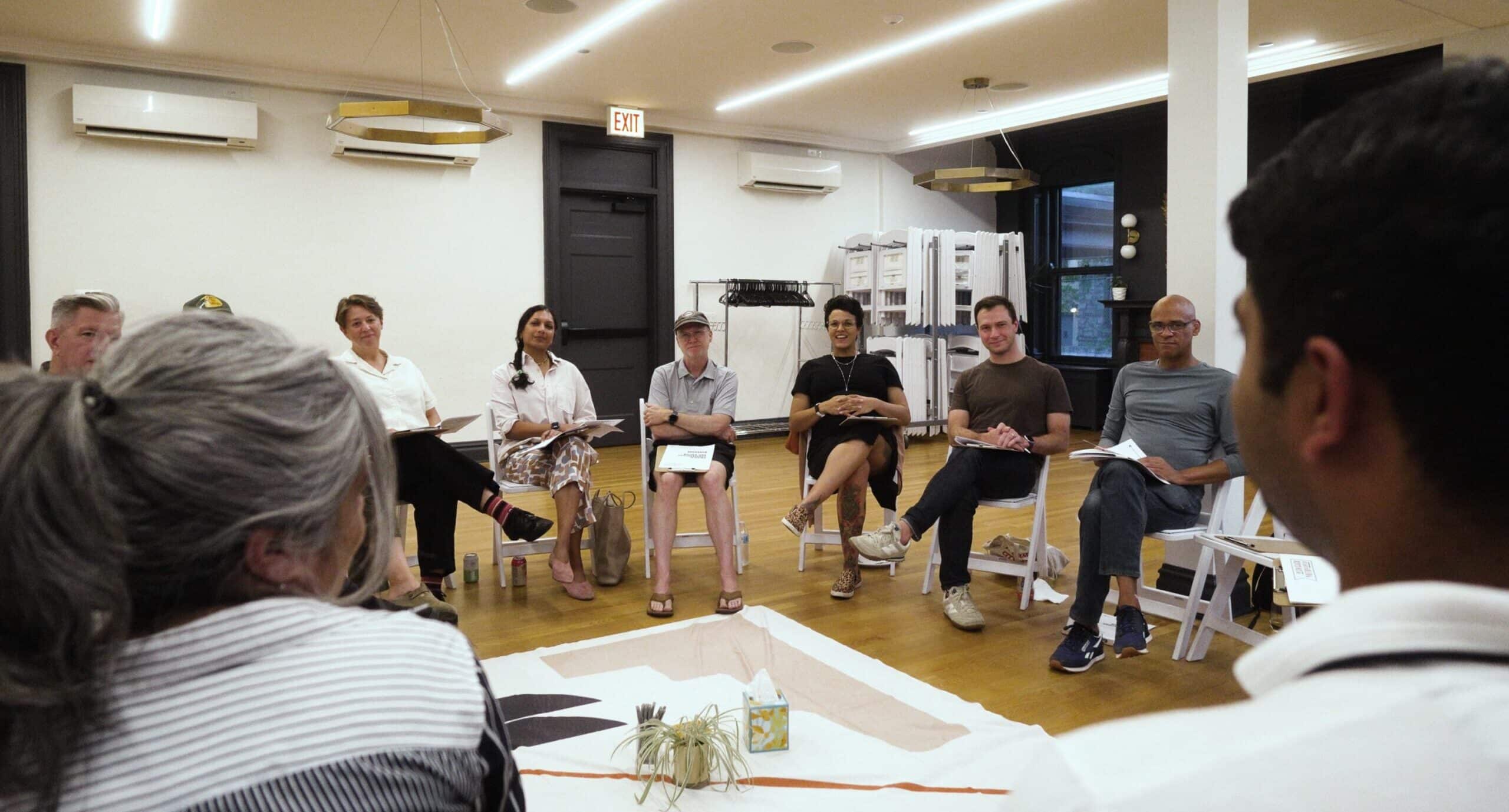 Participants sit in a circle on white folding chairs during a Critical Conversations workshop, with a decorative cloth and small plant arranged in the center of the room.