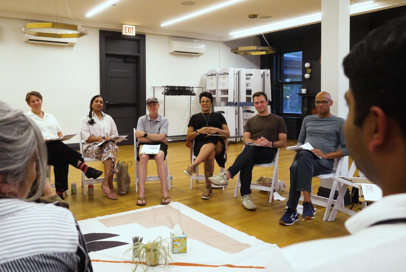 Participants sit in a circle on white folding chairs during a Critical Conversations workshop, with a decorative cloth and small plant arranged in the center of the room.
