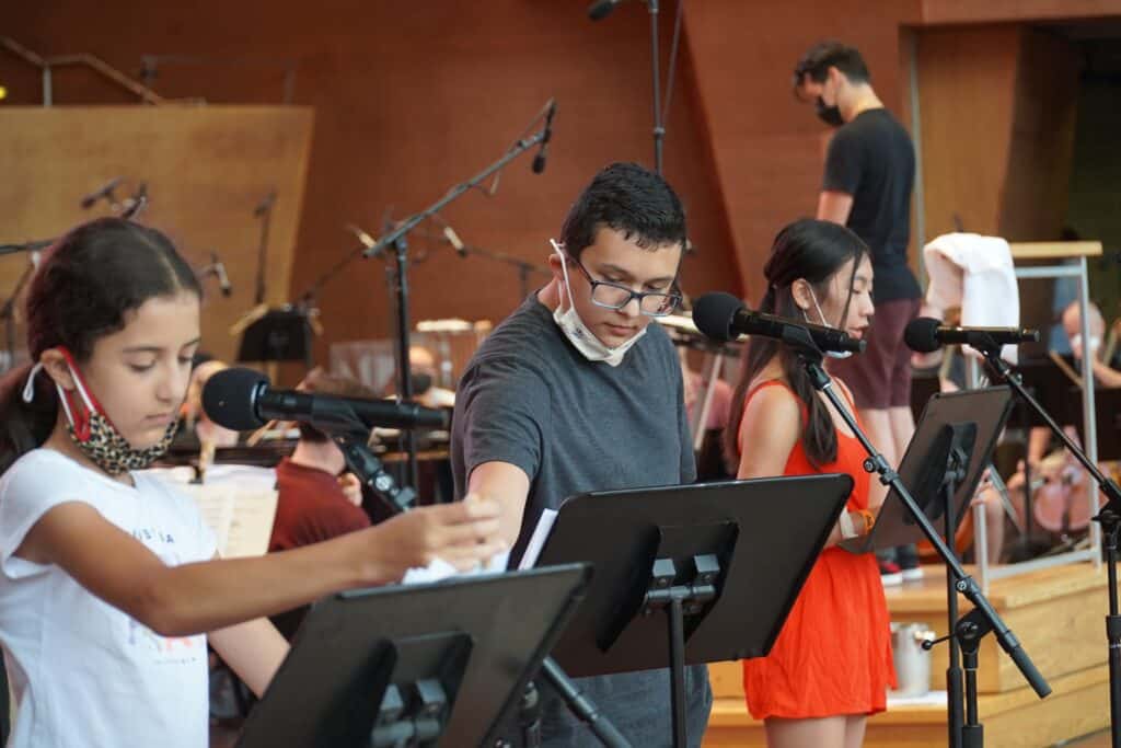 Three young students stand at music stands with microphones, reading their poems aloud on a concert hall stage during a Chicago Poetry Center event.