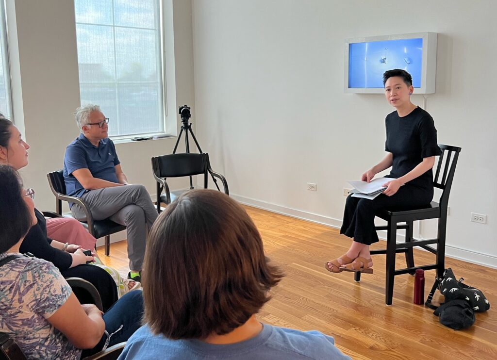 A poet in all black sits in a chair holding papers, speaking to a small audience in a gallery space with a video artwork visible on the wall behind her.