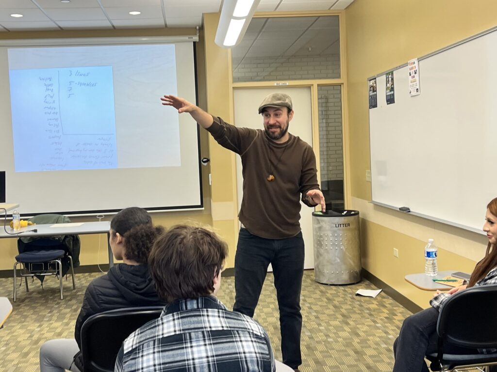 A teaching artist in a flat cap and brown sweater gestures expressively while leading a poetry workshop, with handwritten notes projected on a screen behind him.
