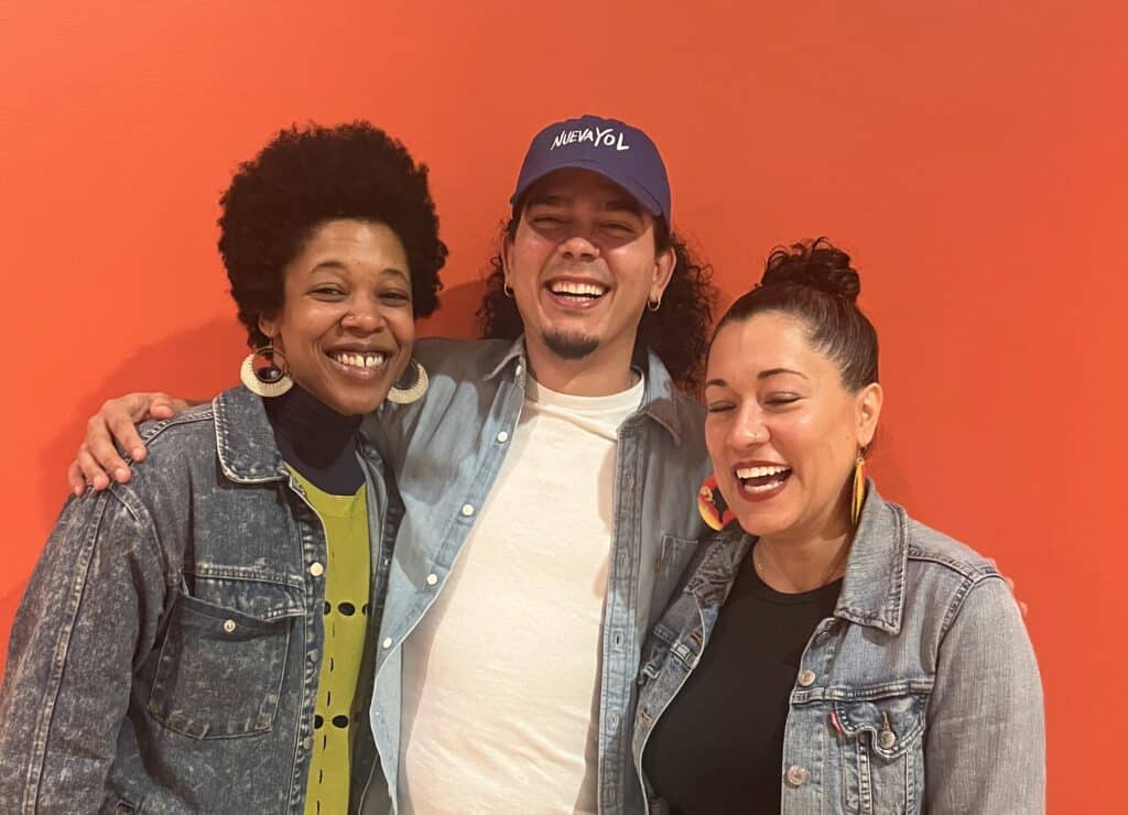 Three people laugh together against a bright orange wall—all wearing denim jackets, with the center person in a ""NuevaYol"" cap—radiating warmth and joy.