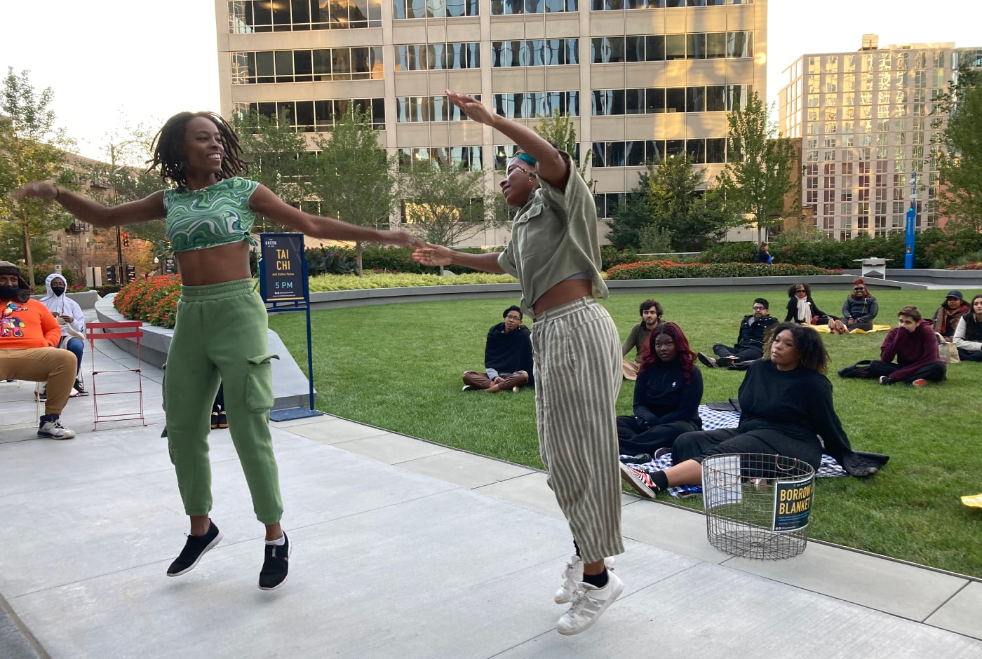 Two dancers leap joyfully on a plaza stage at an outdoor Poetry at The Green event, with audience members seated on the grass and downtown buildings in the background.