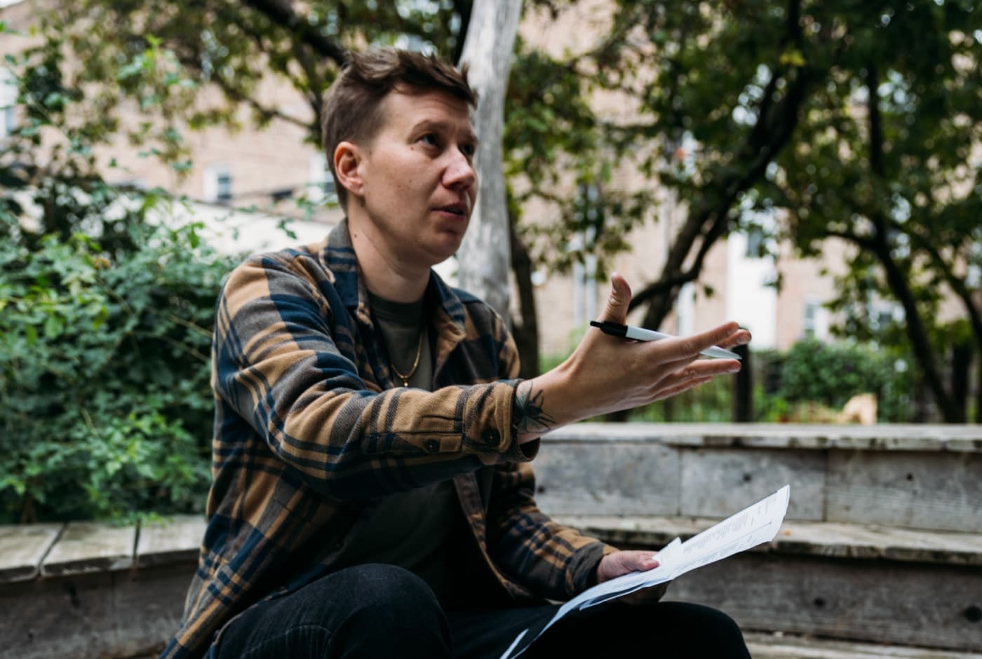 A person in a plaid flannel jacket gestures while holding papers, leading an outdoor poetry discussion on a wooden bench surrounded by greenery.
