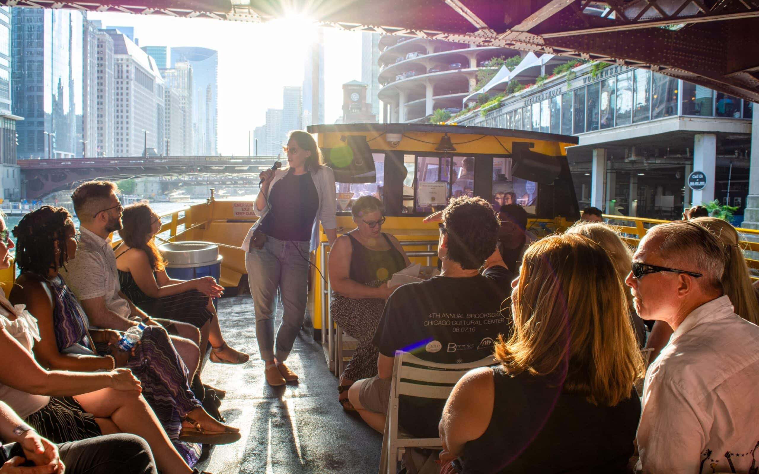 A poet reads into a handheld microphone aboard a water taxi as sunlight streams beneath a bridge, with Chicago's Marina City towers and downtown skyline in the background while audience members listen.