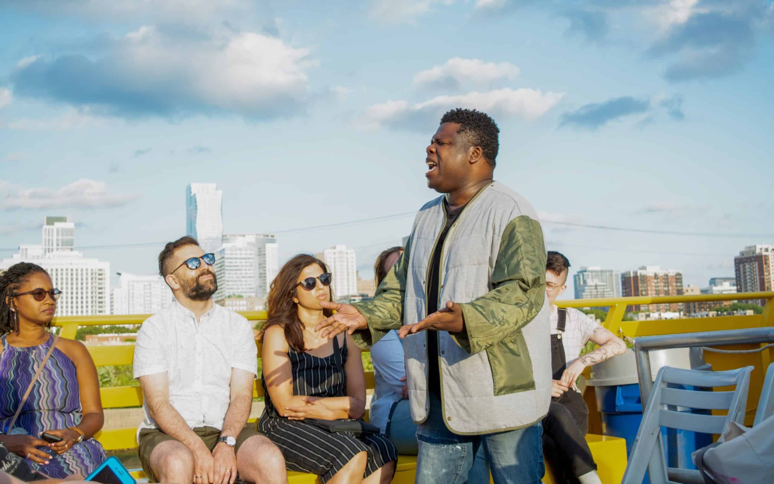 A poet performs on the deck of a boat with the Chicago skyline in the background, gesturing expressively while audience members in sunglasses listen from yellow benches.