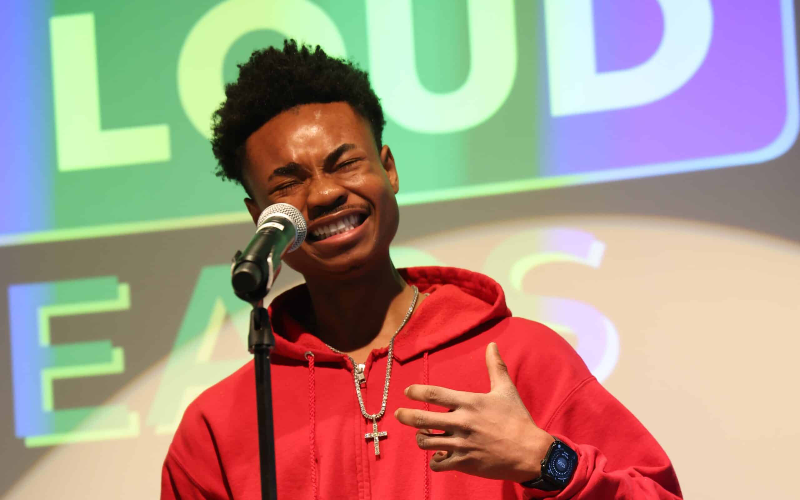 A young man in a red hoodie performs passionately at a microphone, eyes squeezed shut and hand on his chest, with colorful Poetry Out Loud branding projected behind him.