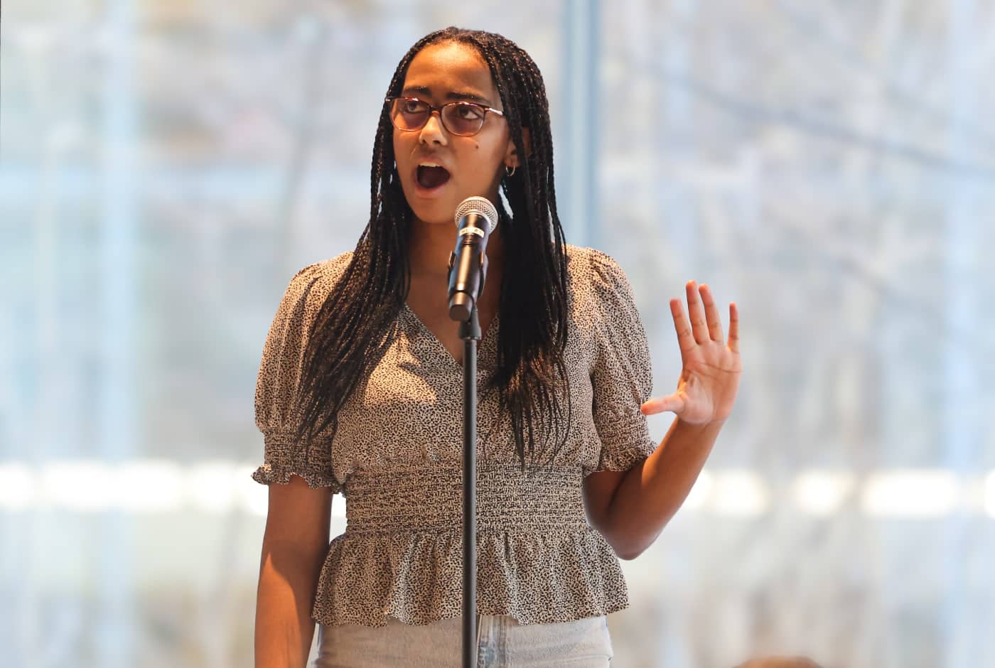 A young woman with long braids and glasses performs poetry at a microphone, one hand raised expressively, wearing a patterned peplum top with soft light behind her.