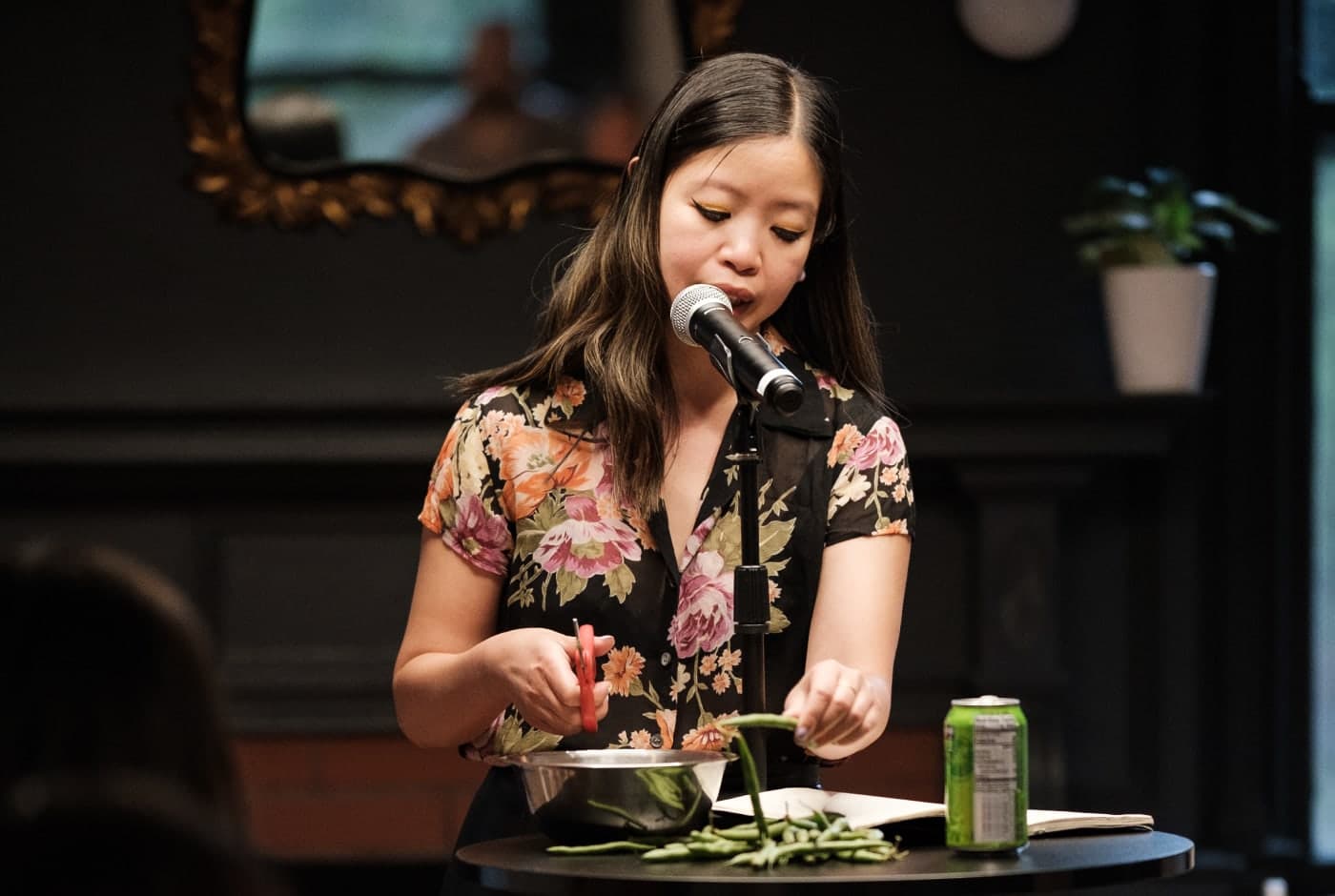 A poet in a floral blouse performs at a microphone while trimming green beans into a bowl—an unconventional, multisensory poetry reading in an intimate venue.