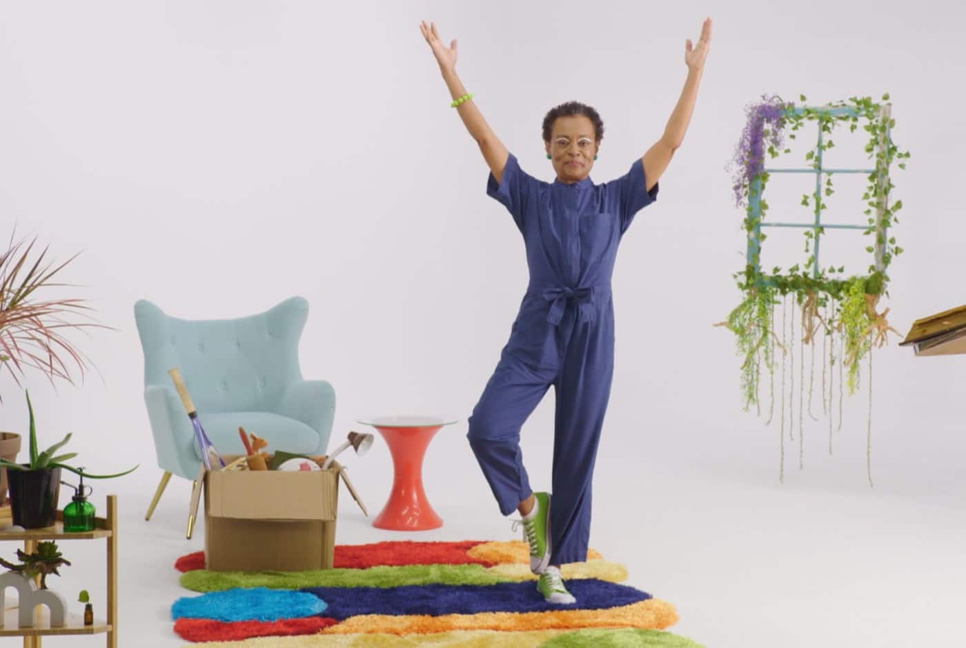 A woman in a blue jumpsuit strikes an exuberant pose with arms raised high on a colorful video set featuring a turquoise armchair, trailing plants, and a rainbow shag rug.