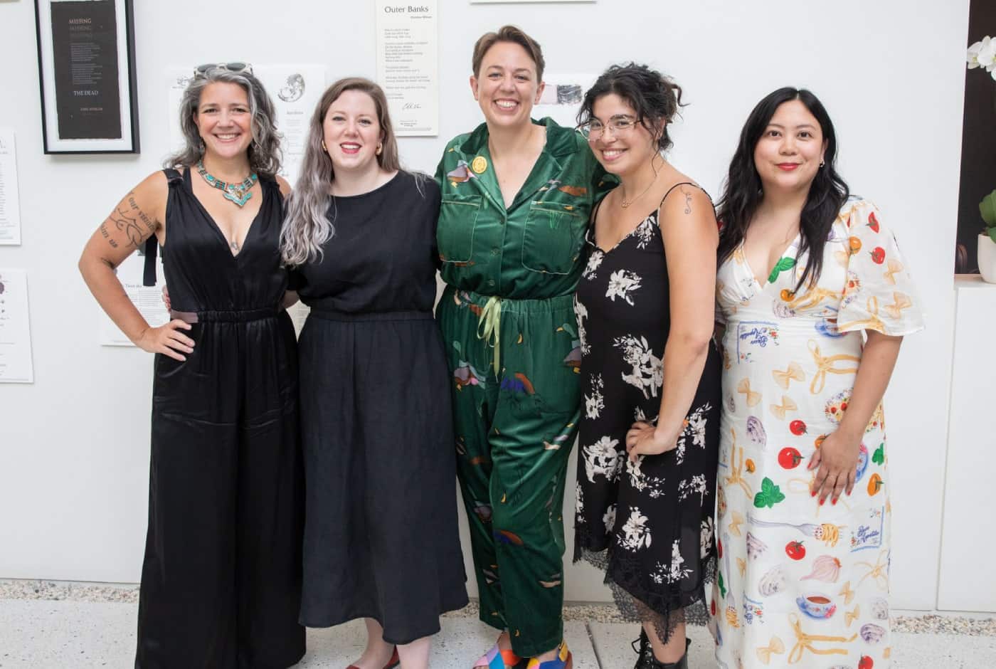 Five women stand together smiling at a gallery event, dressed in elegant attire ranging from black dresses to a green jumpsuit and a playful food-print dress, with poetry broadsheets displayed on the wall behind them.