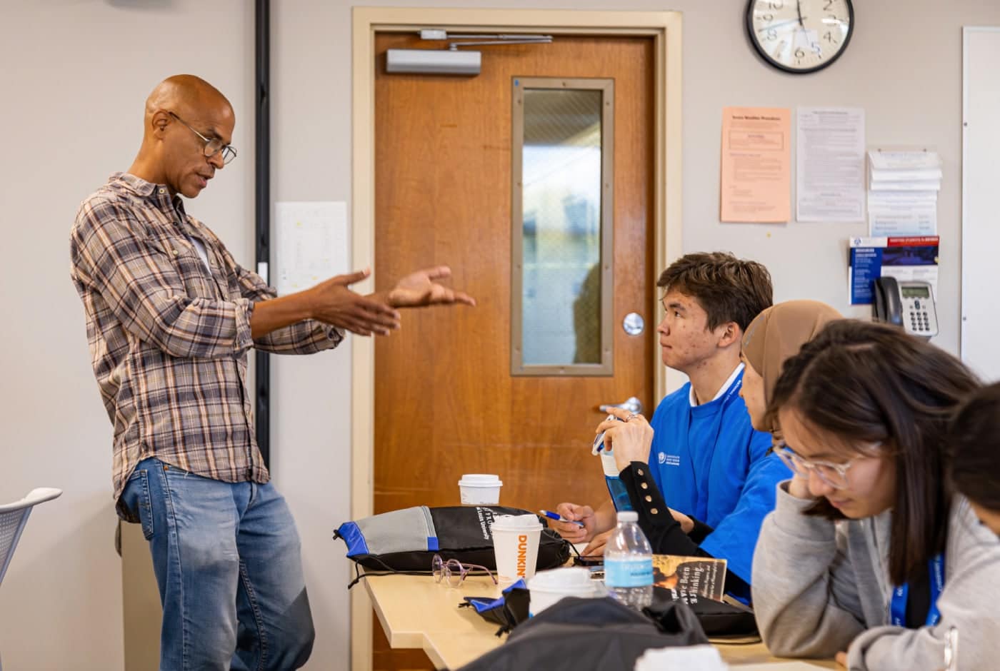 A teaching artist in a plaid shirt gestures while leading a poetry workshop as students at desks listen attentively and take notes.