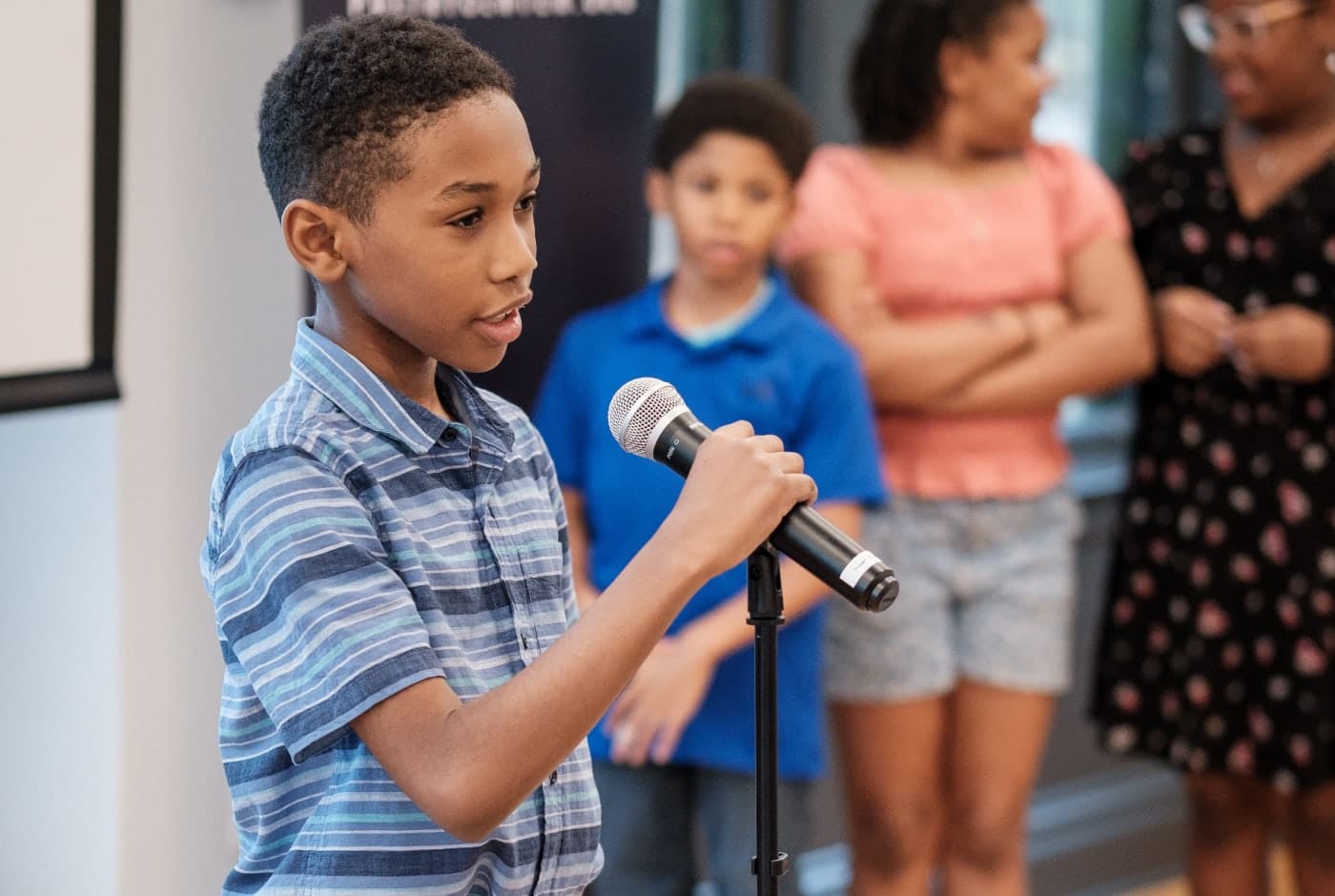 A young boy in a blue striped polo shirt speaks confidently into a handheld microphone at a poetry reading, with other children and an adult waiting nearby.