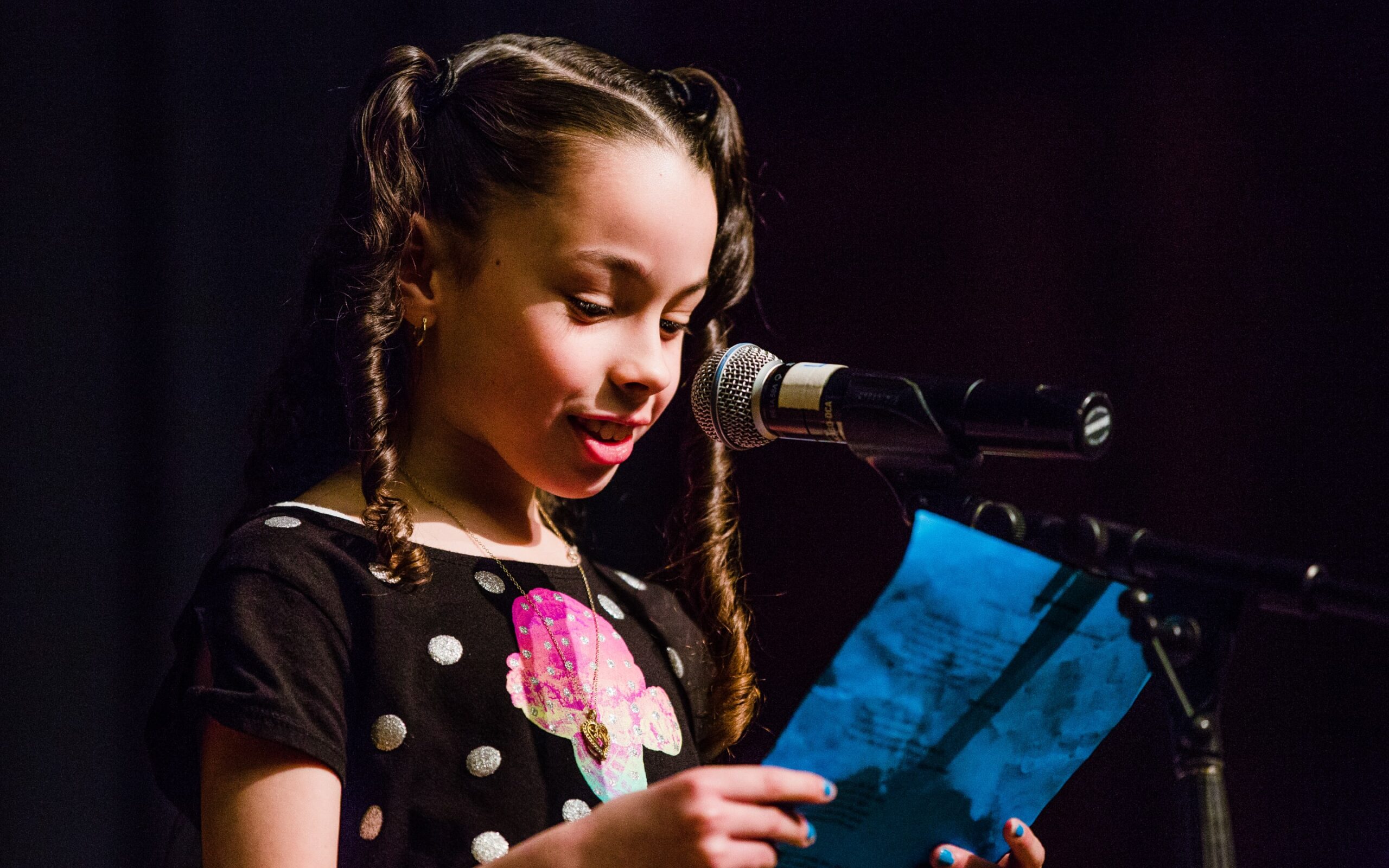 A young girl with braided pigtails reads her poem from a blue paper at a microphone, wearing a black polka-dot top with a colorful mushroom design, with dramatic stage lighting against a dark backdrop.